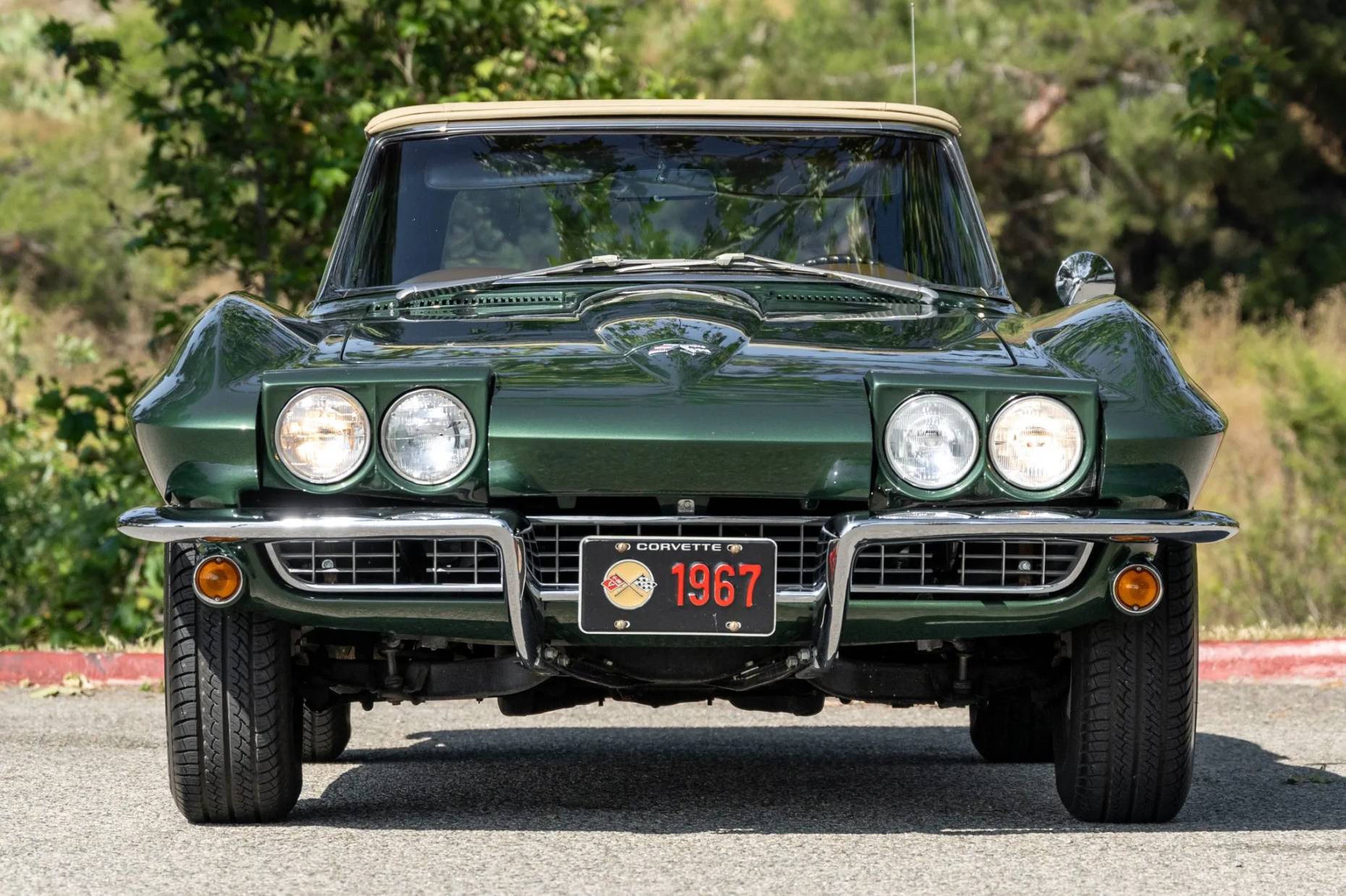 Front view of a green 1967 Chevrolet Corvette convertible with the top up and the headlamps raised; there are trees in the background